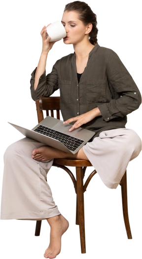 Front view of a young woman wearing home clothes sitting on a chair with a laptop & drinking coffee