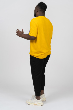 Vista traseira de três quartos de um jovem gesticulando de pele escura em uma camiseta amarela explicando algo