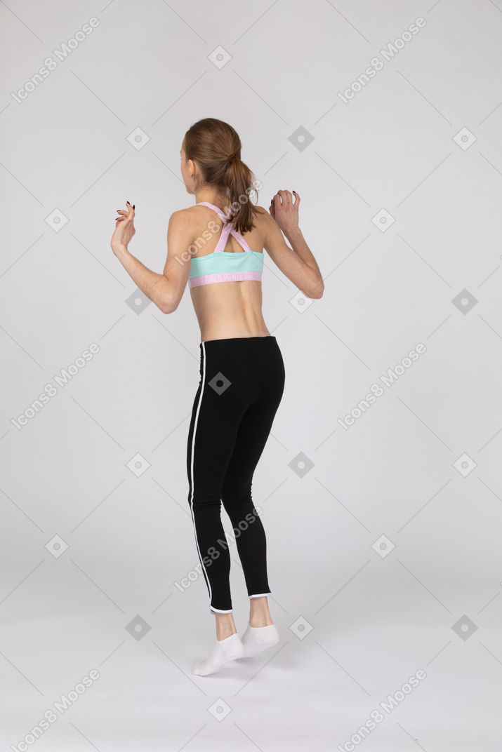 Vista posterior de tres cuartos de una jovencita en ropa deportiva levantando las manos mientras salta