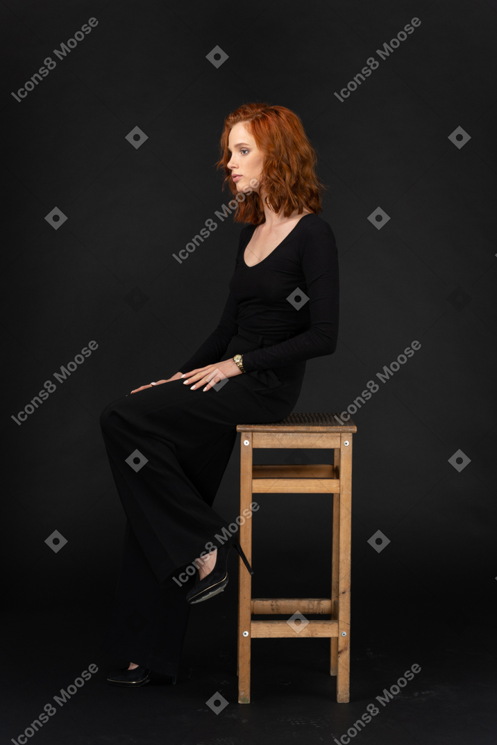 A side view of the beautiful girl sitting on the high wooden chair and holding her hands on knees
