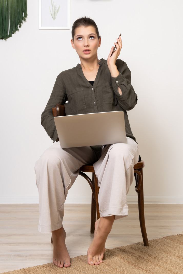 Front view of a shocked young woman sitting on a chair with a laptop & mobile