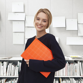 A woman standing in front of a bookshelf holding an orange folder