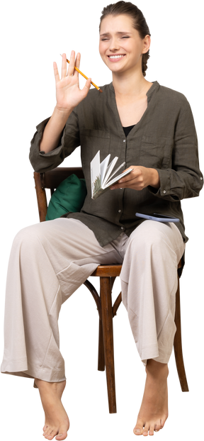 一个微笑的年轻女子穿着家居服坐在椅子上，拿着铅笔和笔记本的前视图