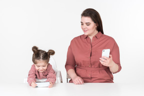 Mãe e sua filha, vestindo roupas vermelhas e rosa, sentadas à mesa de jantar com smartphones nas mãos