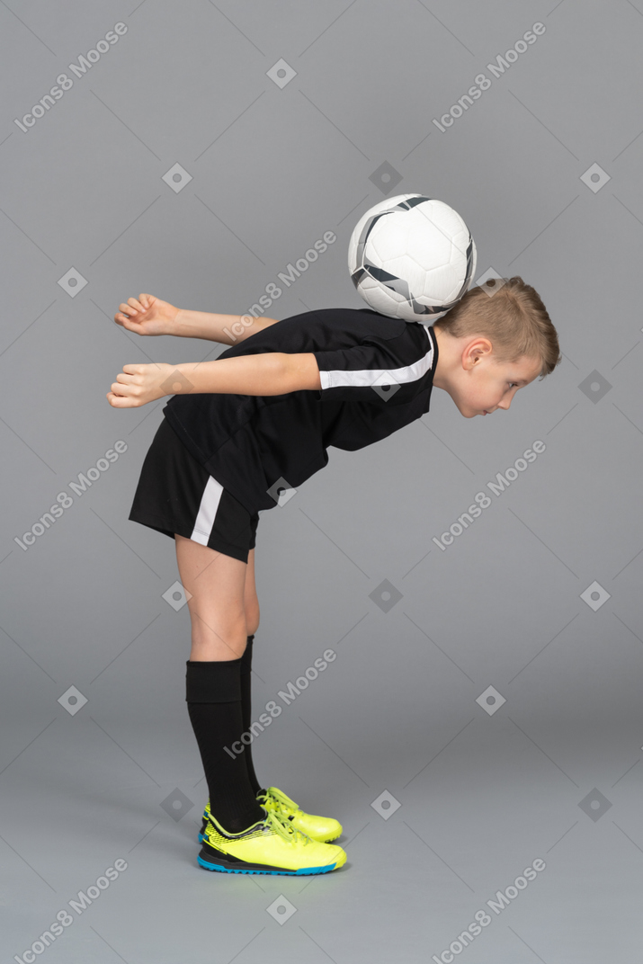 Side view of a child boy in football uniform leaning forward and holding ball on back
