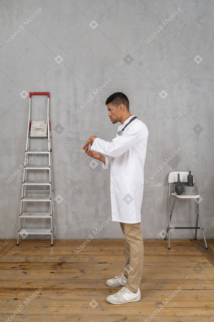 Вид сбоку на молодого врача, стоящего в комнате с лестницей и стулом и держащего руки вместе